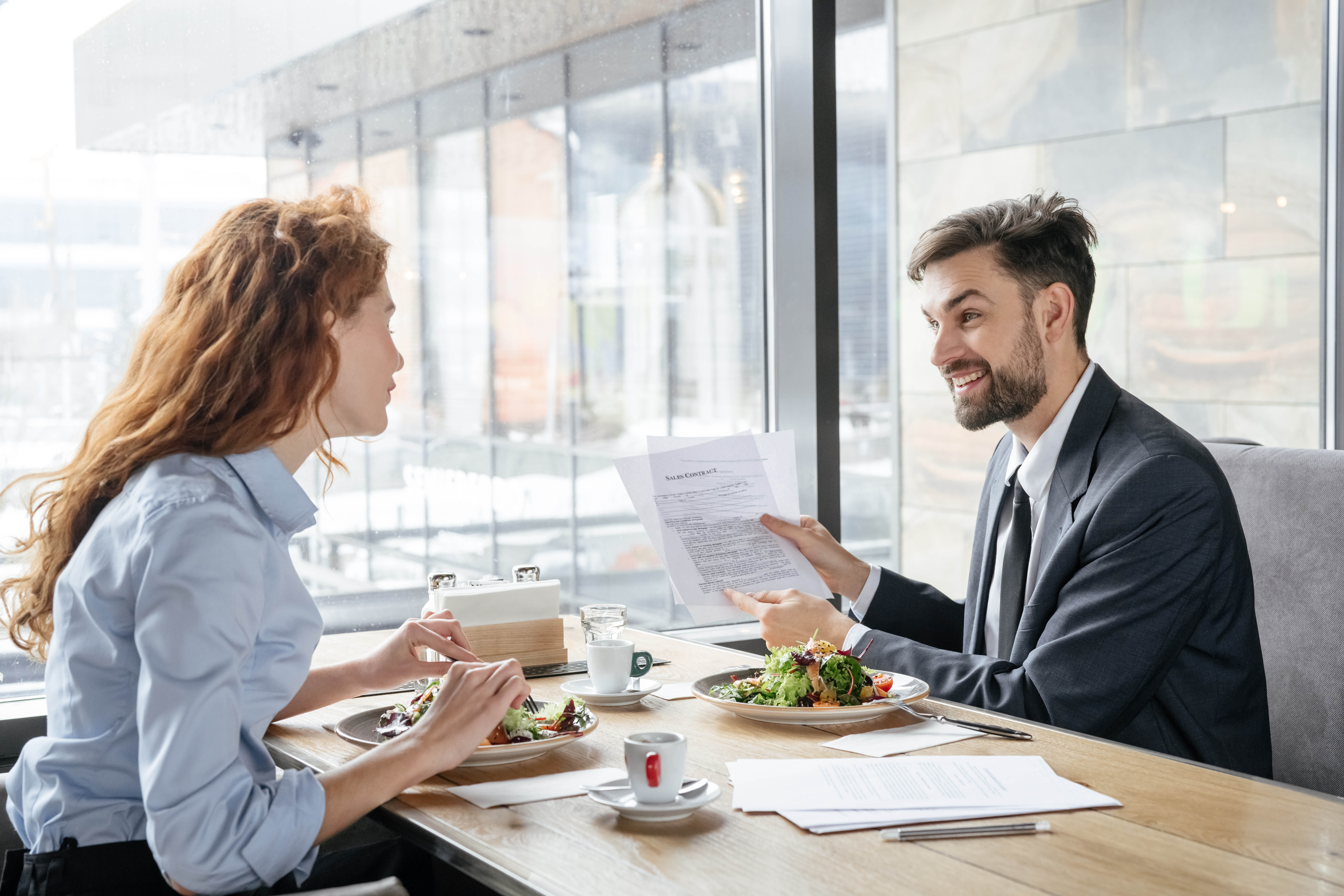 Young man and woman are having a business lunch and reviewing papers that he's holding up, while eating their salads